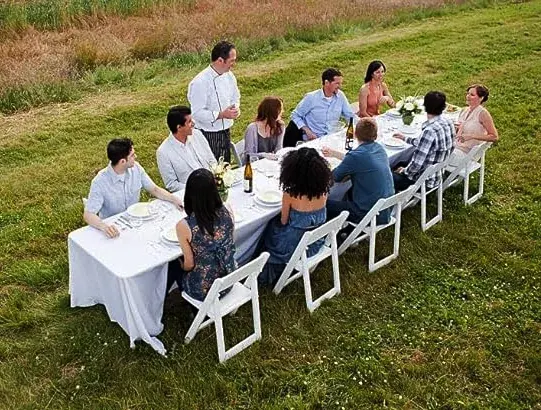 Long banquet table set for outdoor dinner in a field with white folding chairs and a white tablecloth