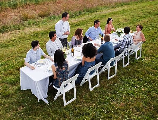 Long banquet table set for outdoor dinner in a field with white folding chairs and a white tablecloth