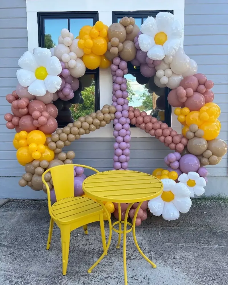 Full circle balloon arch in a daisy peace-sign design with white daisy foils, mustard yellow, mauve, lavender, dusty rose, and tan organic balloons over a yellow bistro chair and table