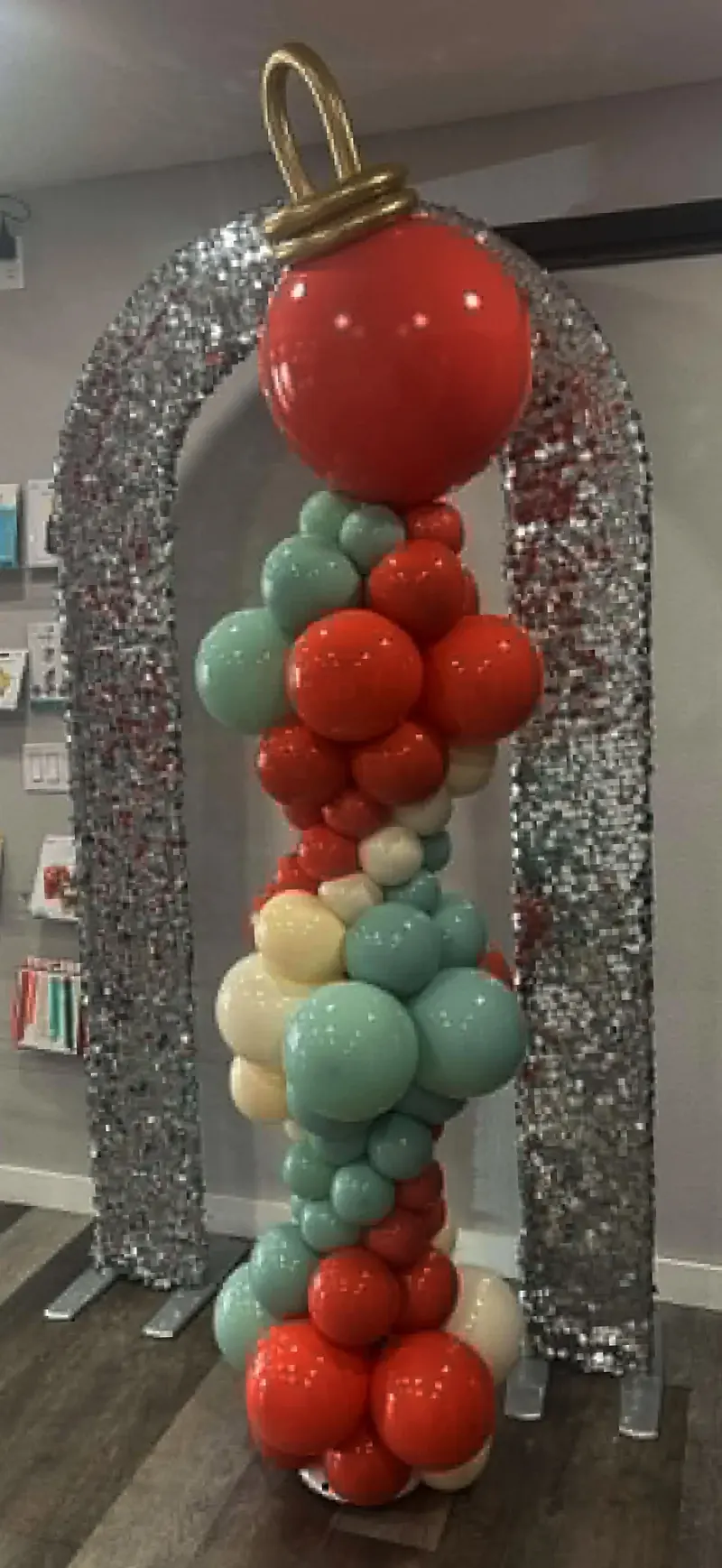 Christmas balloon column in red, teal, and cream with giant red ornament topper inside a silver sequin arch