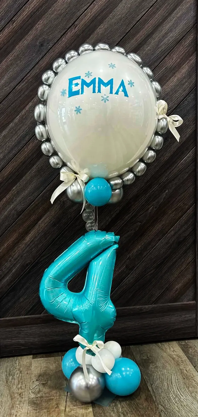 Frozen-themed personalized centerpiece with a clear bubble balloon ringed in chrome silver, custom 'EMMA' lettering and snowflakes, topped over a teal number 4 foil with a satin bow and teal, silver, and white balloon base