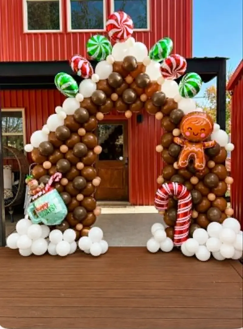 Gingerbread house balloon arch in brown, tan, and cream with peppermint candy foils, red and green candy spinners, a smiling gingerbread man foil, a candy cane foil, and a Happy Holidays mug foil on a red barn porch
