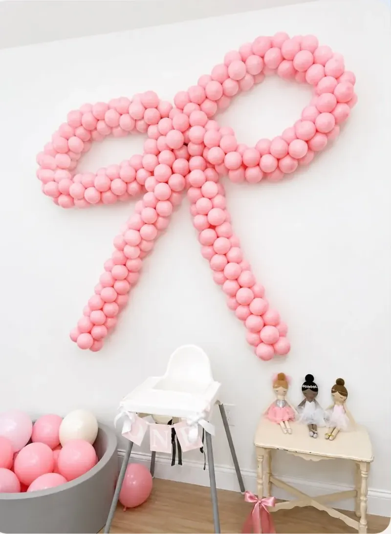 Oversized pink balloon bow sculpture mounted on a white wall above a high chair and ballerina dolls for a first birthday