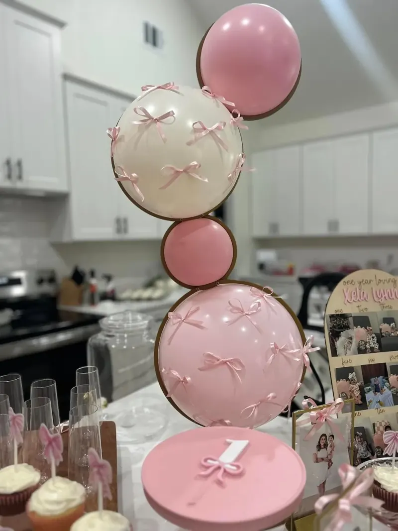 Stacked stuffed-balloon centerpiece with pink and cream orbs tied with pink satin bows on a pink cake plinth for Xela Lynn's first birthday