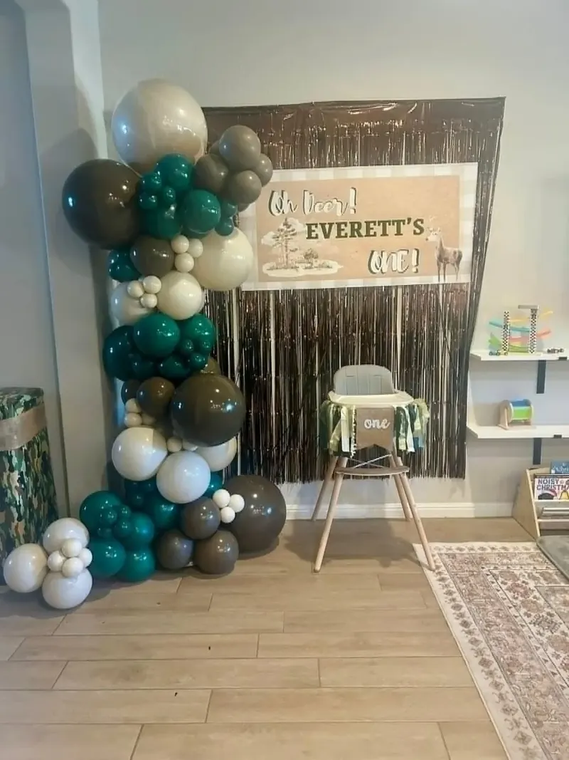 Free-standing 'Oh Deer Everett's One' woodland first birthday balloon column in forest green, brown, and cream with a metallic brown fringe backdrop and high chair