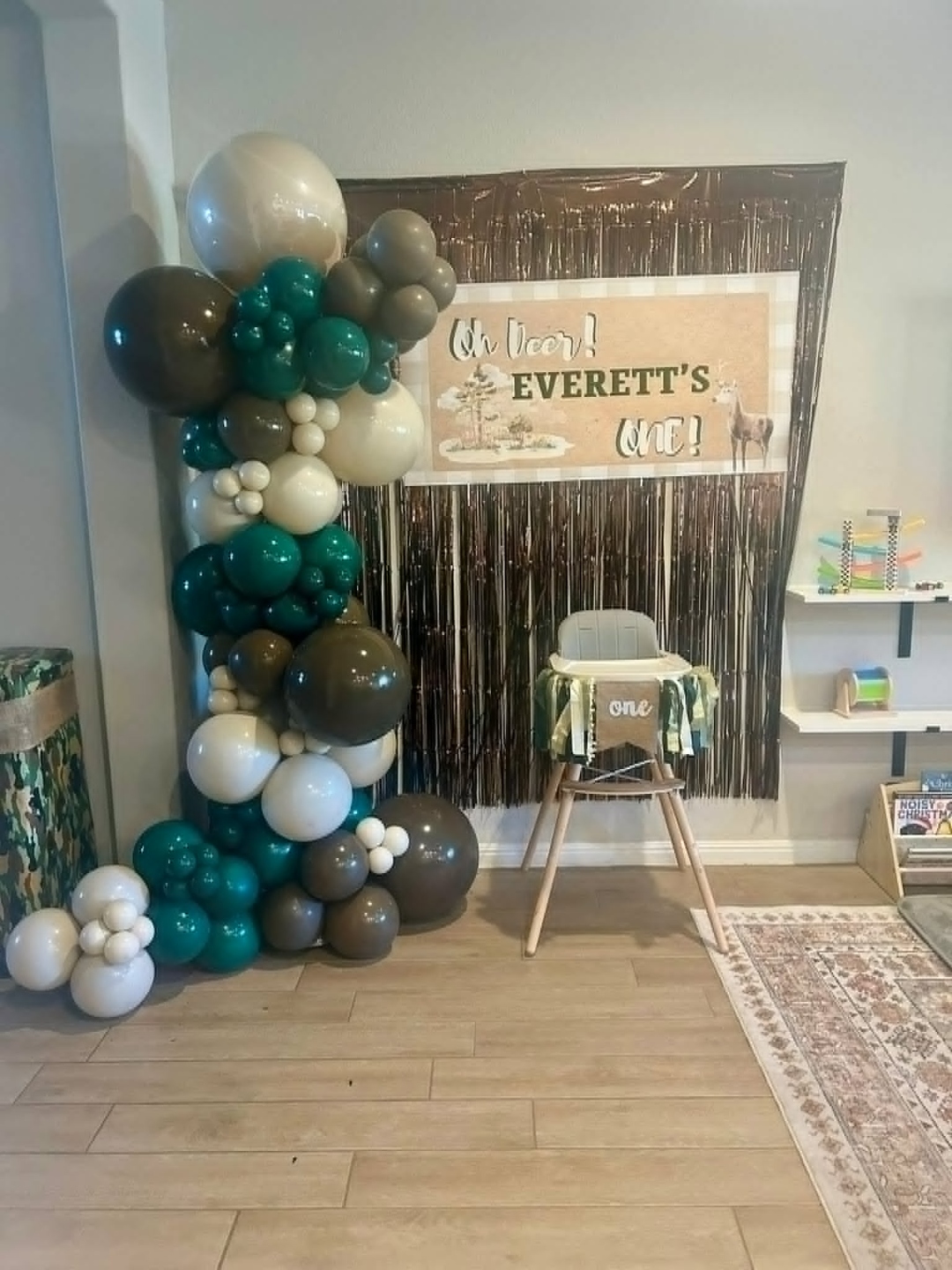 Free-standing 'Oh Deer Everett's One' woodland first birthday balloon column in forest green, brown, and cream with a metallic brown fringe backdrop and high chair