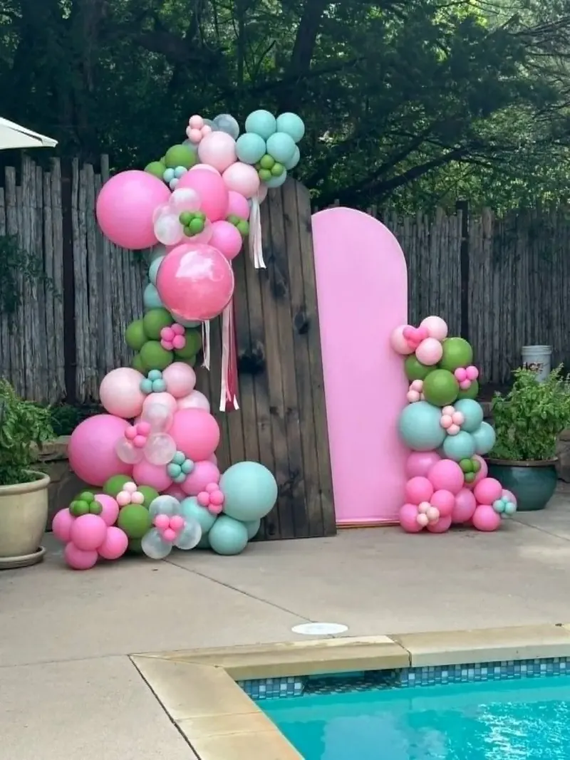 Poolside balloon garland on pink and dark wood arch backdrops in pink, mint, green, and clear balloons with flower clusters and ribbon streamers