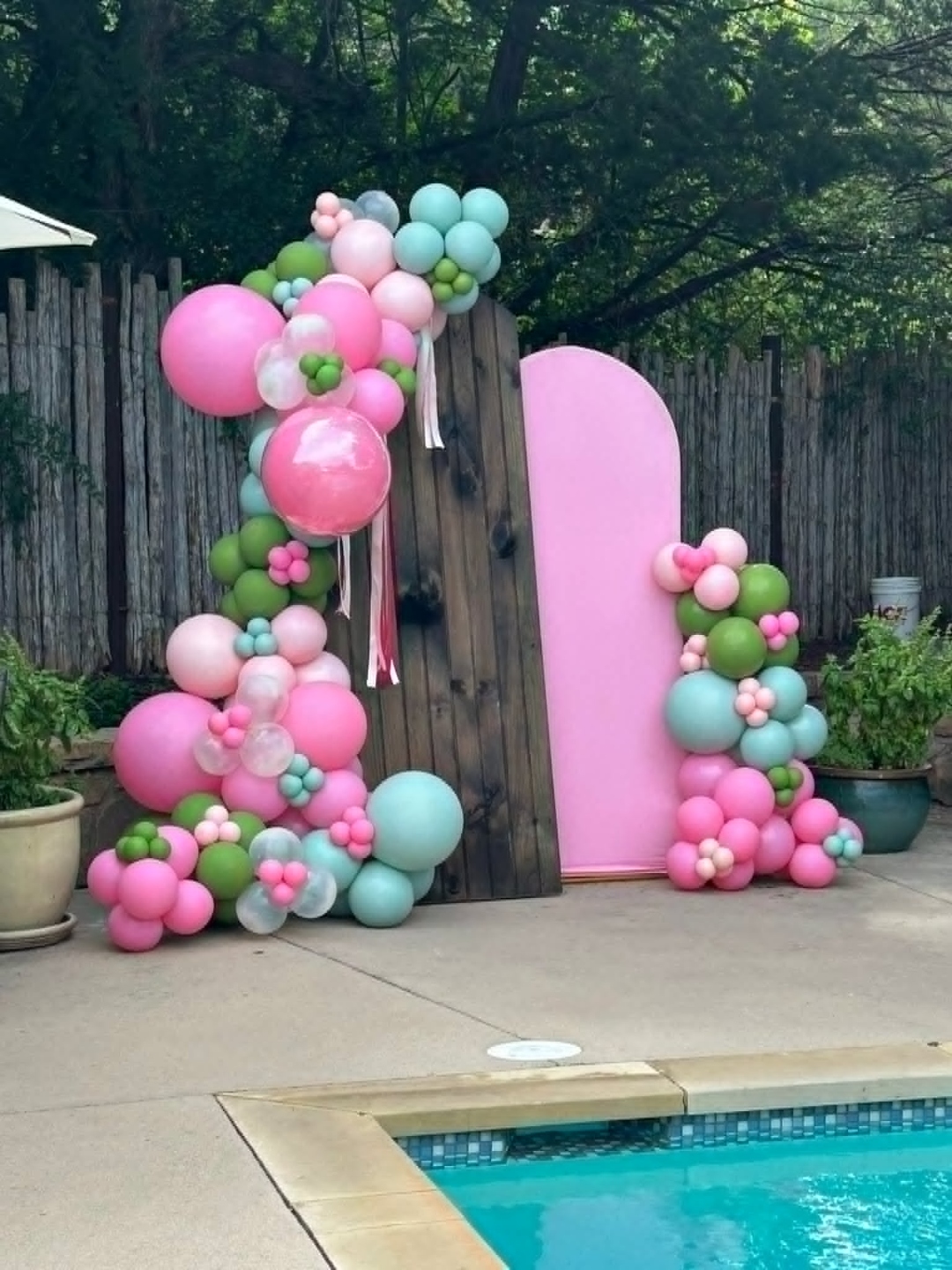 Poolside balloon garland on pink and dark wood arch backdrops in pink, mint, green, and clear balloons with flower clusters and ribbon streamers