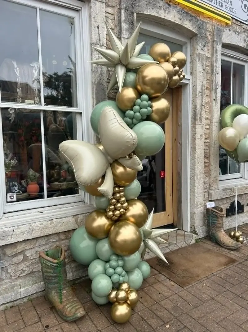 Storefront balloon garland in sage green, chrome gold, and cream with starburst foils, sage grape clusters, and a tall cream leaf foil