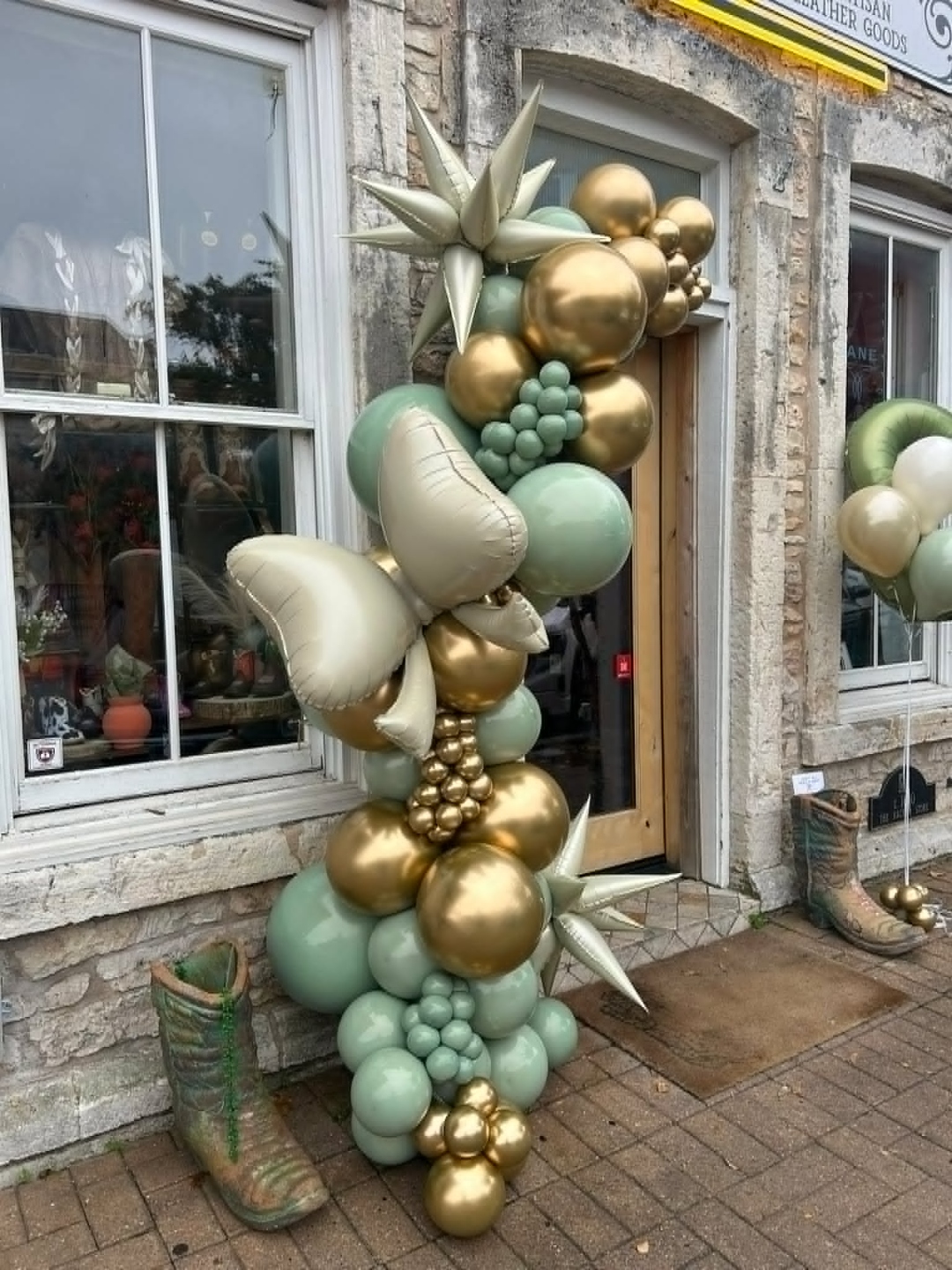 Storefront balloon garland in sage green, chrome gold, and cream with starburst foils, sage grape clusters, and a tall cream leaf foil