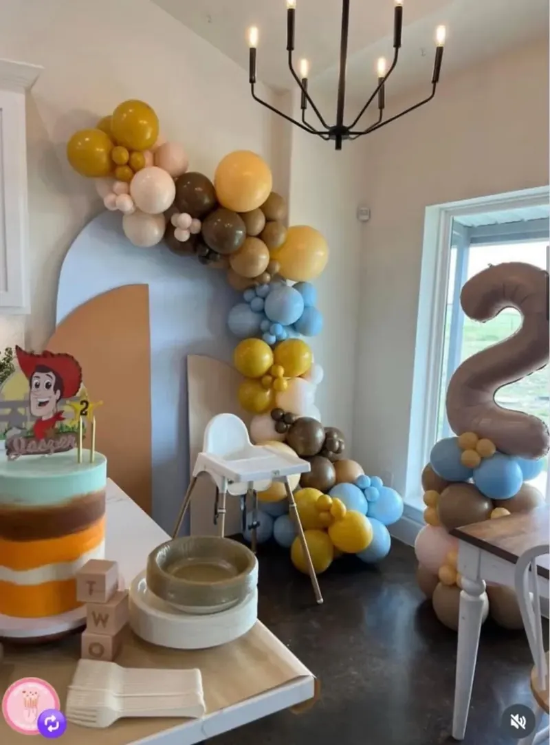 Toy Story 2nd birthday balloon garland in mustard, cream, brown, blue, and yellow draped over a blue arched backdrop with brown number 2 foil column