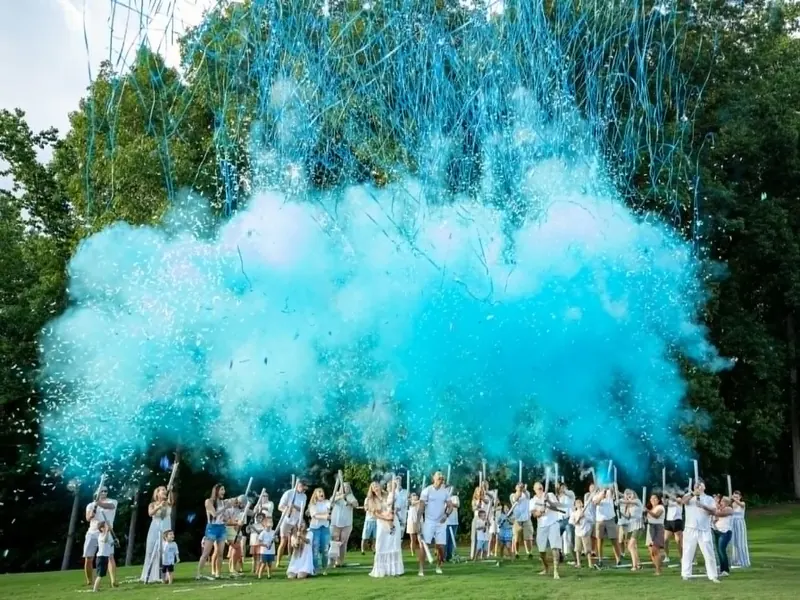 Outdoor gender reveal with a large crowd firing blue powder cannons and streamers, creating a giant cloud of blue smoke and confetti above a grassy field