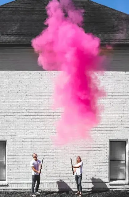 Couple firing pink smoke cannons against a white brick wall, sending a tall plume of pink smoke into the air for a gender reveal