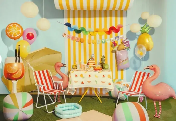 Summer luau party scene with flamingo balloons, orange slice and pineapple foils, paper lanterns, and a birthday cake under yellow and white stripes