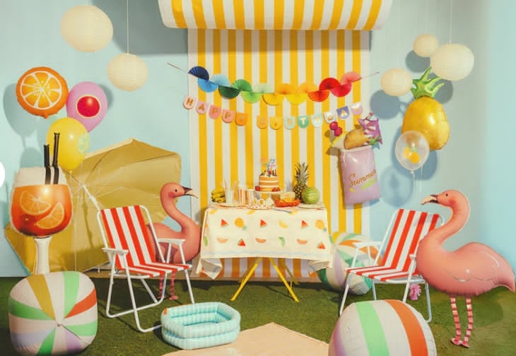 Summer luau party scene with flamingo balloons, orange slice and pineapple foils, paper lanterns, and a birthday cake under yellow and white stripes
