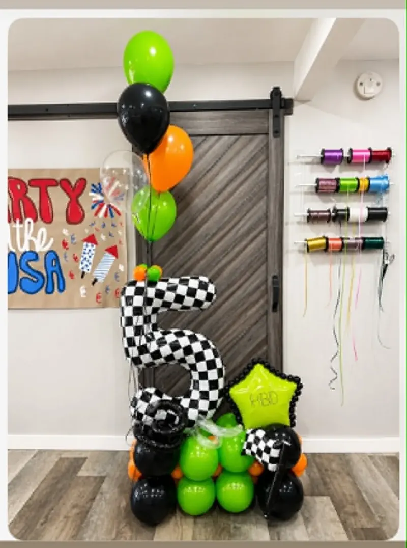 Racing-theme 5th birthday balloon stack with checkered flag number 5, green HBD star foil, and lime green, orange, and black balloon base