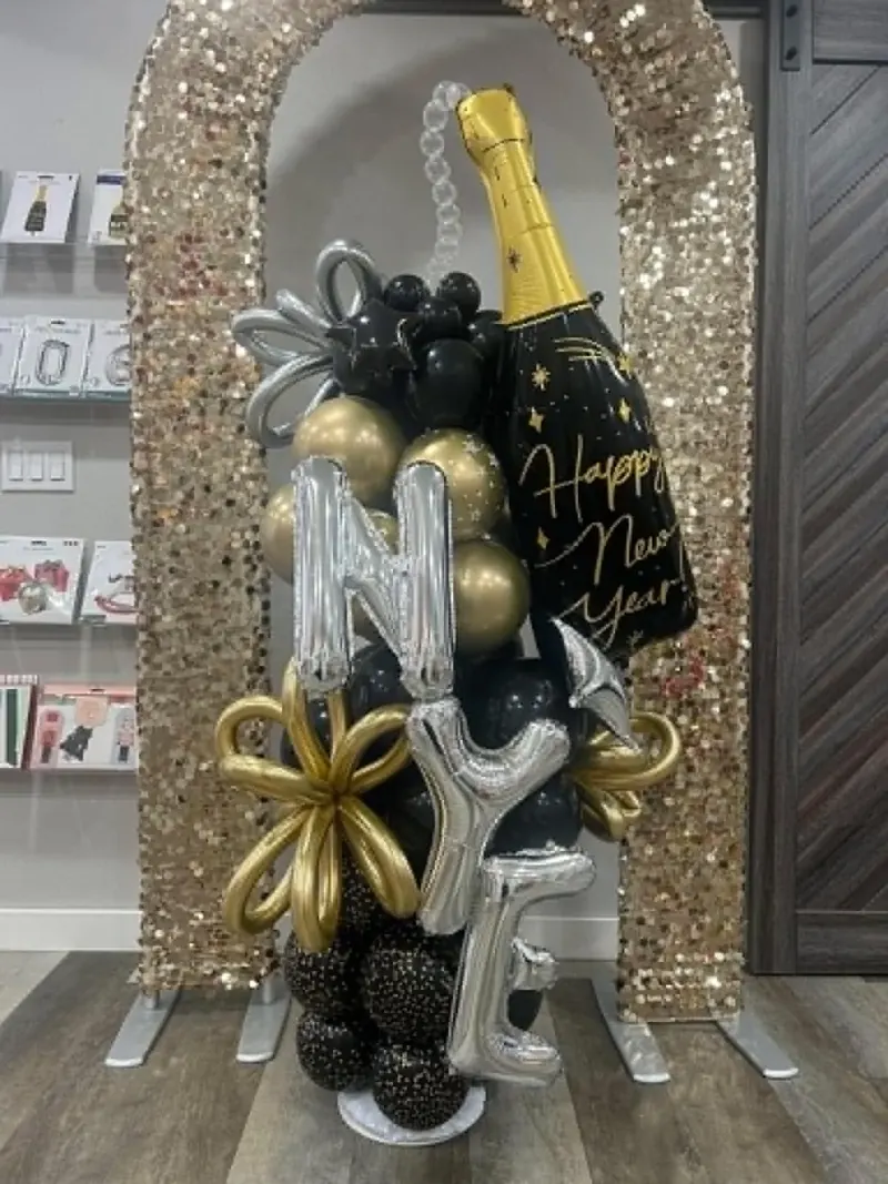 Happy New Year balloon stack with gold and black champagne bottle foil, silver NYE letter balloons, and gold flower accents under a gold sequin arch