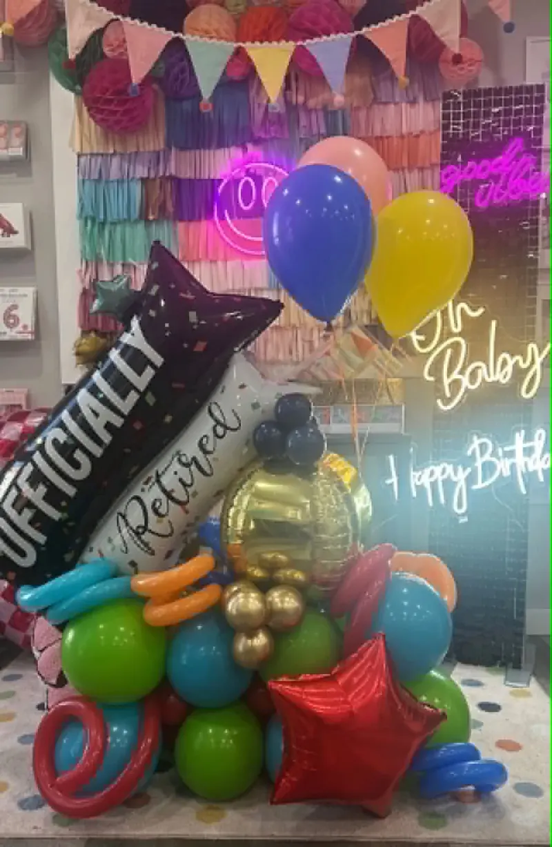 Officially Retired balloon stack with black confetti star foil, white retired sash, gold orb, blue and yellow round balloons, red star, and rainbow base of green, blue, orange, and pink balloons