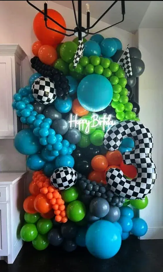 Racing-theme balloon wall in teal, lime, black, and orange with checkered foil flags and 'Happy Birthday' neon sign